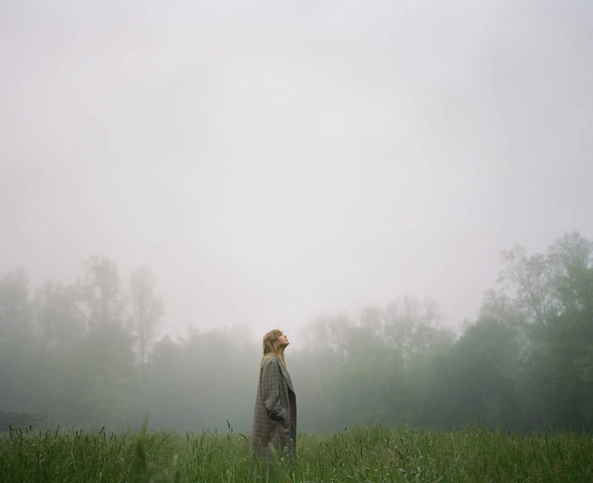 Taylor Swift standing in a misty field wearing a checked coat.