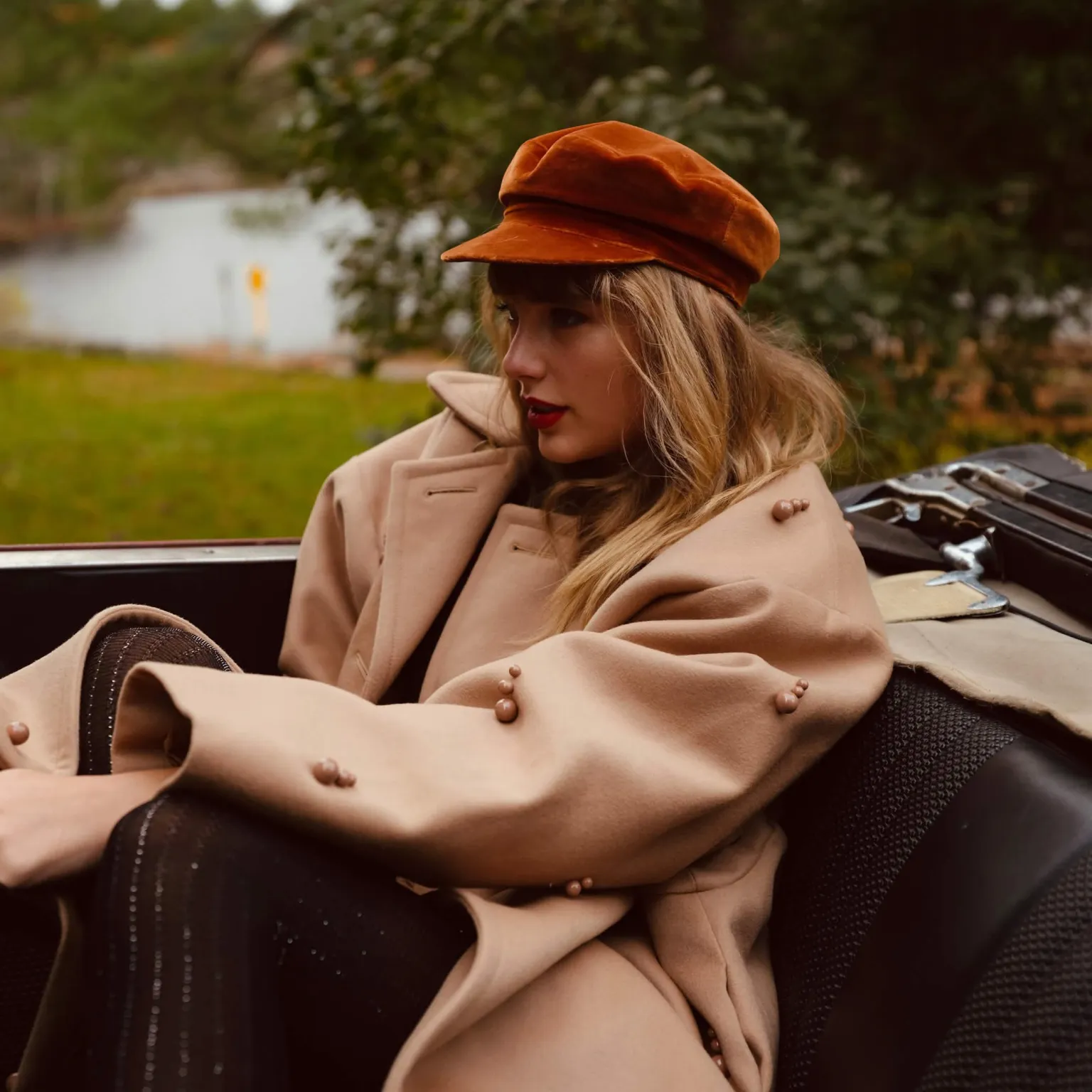 Taylor Swift wearing a red cap and beige coat in a convertible.
